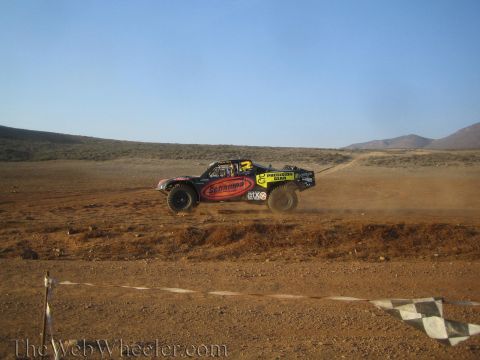 1990 Ford Bronco - black and yellow off-road race truck with Schampa and Precision Gear decals races across dry desert scrub past a checkered marker.