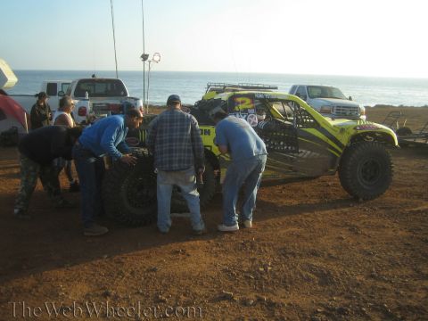 1990 Ford Bronco - pit crew gathers around a yellow-green off-road race truck on a coastal bluff at dusk, with the ocean and support trucks behind.