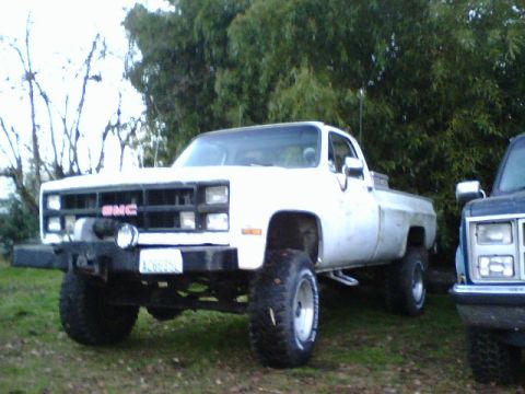 1987 Ford Bronco - A white GMC pickup truck is parked outdoors next to another vehicle, with trees in the background.