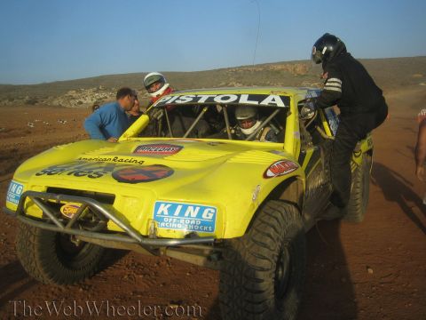1990 Ford Bronco - Crew members lean over a yellow off-road race truck lettered Pistola while a helmeted driver sits inside, on a desert track at sunset.