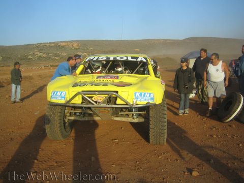 1990 Ford Bronco - Front view of a yellow off-road race truck lettered Pistola surrounded by spectators and crew on a dirt desert course.