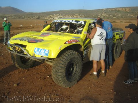 1990 Ford Bronco - Crew members service a yellow off-road race truck lettered Pistola on a dirt desert track, with onlookers and another race truck behind.