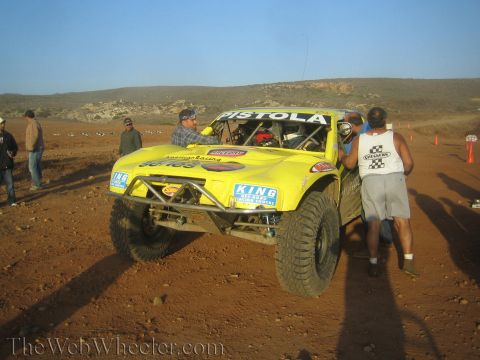 1990 Ford Bronco - yellow off-road race truck lettered Pistola sits on a dirt desert track at golden hour as crew and spectators gather around it.