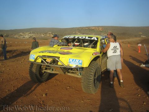 1990 Ford Bronco - yellow off-road race truck lettered Pistola is attended by a crew member on a dusty desert course under a low sun.
