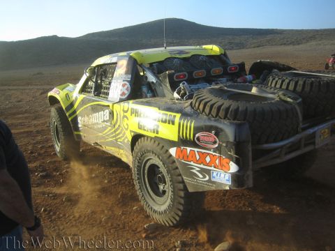 1990 Ford Bronco - Rear quarter view of a green and yellow off-road race truck carrying spare tires with Schampa and Maxxis decals, kicking dust in the desert.