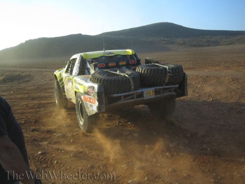 1990 Ford Bronco - green and yellow off-road race truck loaded with spare tires drives away into the desert toward a hazy low sun, trailing dust.