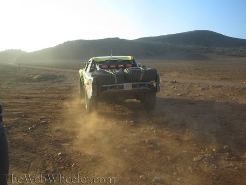 1990 Ford Bronco - off-road race truck carrying spare tires drives away across rocky desert ground toward a bright low sun, raising dust.