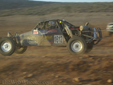1990 Ford Bronco - open tube-frame off-road race buggy numbered 1612 races across rocky desert terrain with a chase vehicle behind.