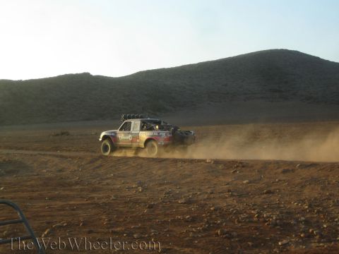 1990 Ford Bronco - off-road race truck with a roof light bar speeds across dusty desert flats below a dark hill, trailing a plume of dust.