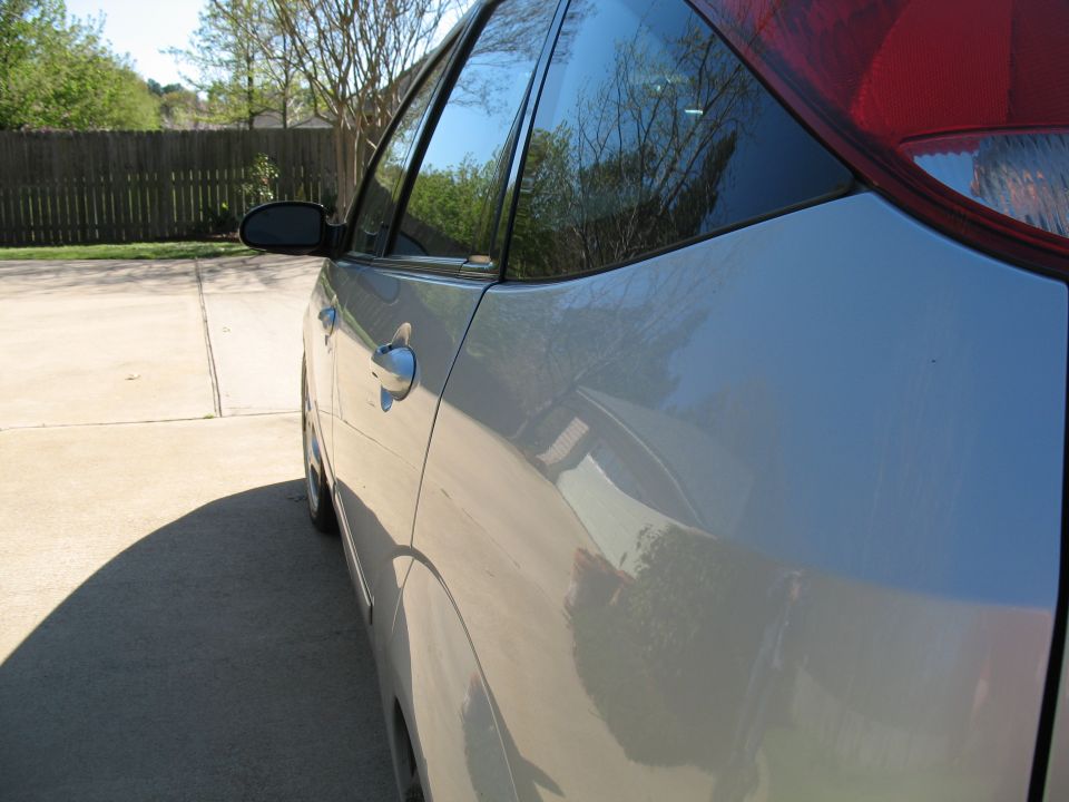 Silver car side profile, showing door handle and rear window. Trees reflected in glass