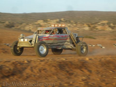 1990 Ford Bronco - gray tube-frame off-road race buggy numbered 1311 with a roof light bar races across reddish desert terrain.