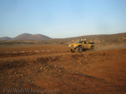 1990 Ford Bronco - yellow off-road race truck with a roof light bar drives a dirt desert trail past course markers, a mountain rising in the distance.