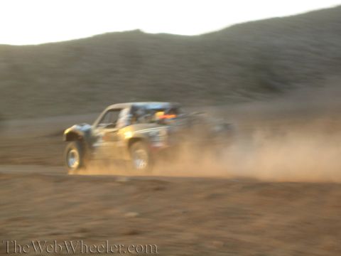 1990 Ford Bronco - motion-blurred off-road race truck speeds through a heavy cloud of dust on a dirt course below hills at dusk.