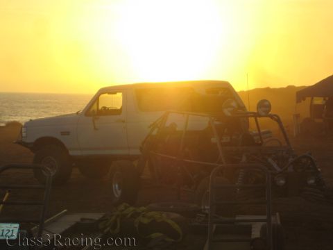 1990 Ford Bronco - white early Ford Bronco and tube-frame race buggies are silhouetted against a brilliant ocean sunset at a coastal pit area.