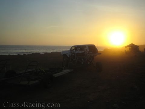 1990 Ford Bronco - tube-chassis off-road race truck sits on a flatbed trailer at a coastal desert pit as the sun sets over the sea.