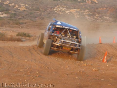1990 Ford Bronco - blue tube-framed off-road race truck carrying number 7 charges through a bermed dirt corner marked by orange cones, throwing up dust.