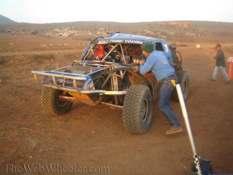 1990 Ford Bronco - crew member in a green beanie fuels a mud-caked blue tube-chassis race truck at a desert pit stop near downed course markers.