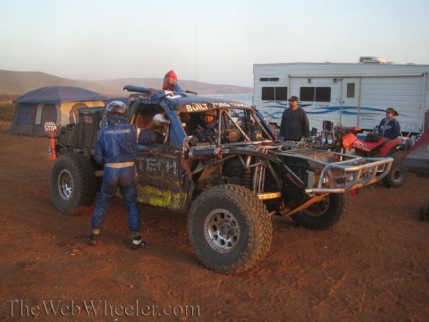 1990 Ford Bronco - Crew members in racing suits service a dusty tube-chassis off-road truck at a dirt pit area beside an RV and tents at dusk.
