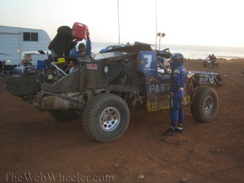 1990 Ford Bronco - blue number 7 tube-chassis race truck is refueled from a red jug at a dirt pit by the ocean while a driver in blue suit stands by.
