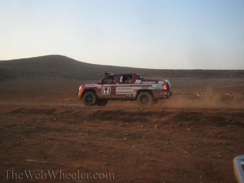 1990 Ford Bronco - red and white Honda-badged crew-cab race truck races side-on across reddish desert dirt, trailing dust at dusk.