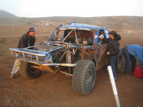 1990 Ford Bronco - Several crew members surround a mud-spattered blue tube-chassis off-road truck during a desert pit stop with red fuel jugs nearby.