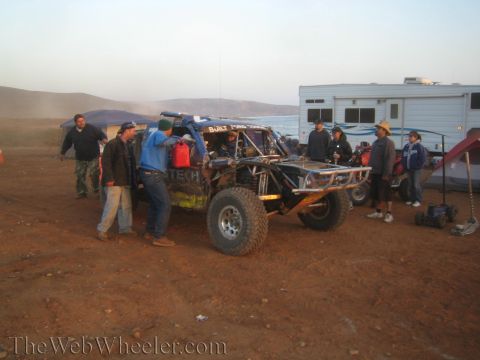 1990 Ford Bronco - group gathers around a tube-chassis off-road race truck at a hazy dawn pit camp with RVs, tents and the sea in the distance.