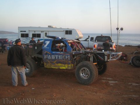 1990 Ford Bronco - blue number 7 'Fastech' tube-chassis race truck sits at a coastal dirt pit flanked by an RV and support trucks at dusk.