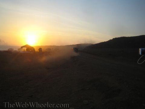 1990 Ford Bronco - Vehicles and figures are silhouetted in a haze of dust at a desert pit area as the sun sets behind the hills.