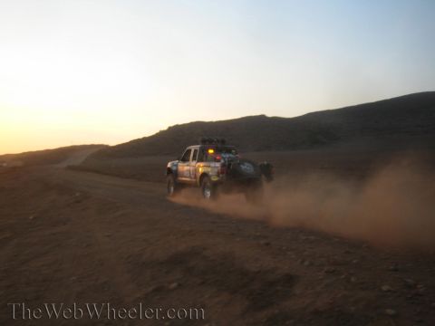 1990 Ford Bronco - off-road race truck with a roof spare tire kicks up a thick dust trail as it heads down a desert track at sunset.