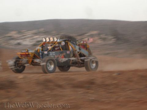 1990 Ford Bronco - colorful open tube-frame sand buggy with a small flag races across reddish desert terrain in a blur of motion.