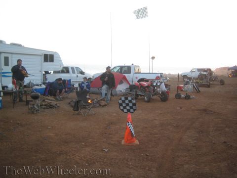 1990 Ford Bronco - dawn desert pit camp shows people by a campfire, RVs and trucks, a red ATV and an orange cone marked with a checkered flag.