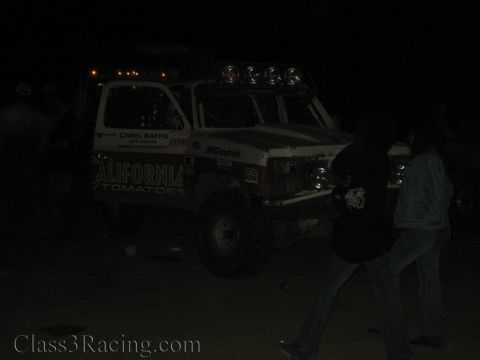 1990 Ford Bronco - white off-road race truck lettered 'California Tomatoes' and 'Chris Raffo' is parked at night as figures walk past in the dark.