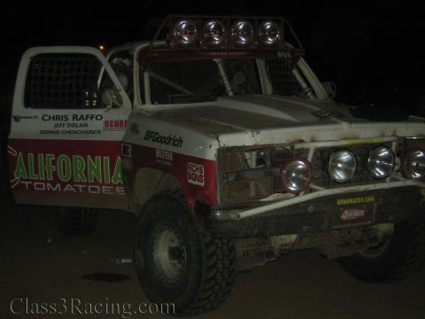 1990 Ford Bronco - Close night view of a white tube-chassis race truck with a roof light bar, BFGoodrich tires and 'California Tomatoes' door livery.