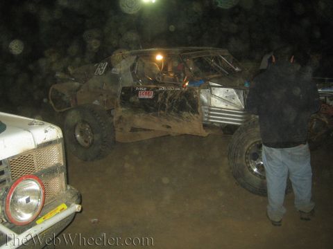 1990 Ford Bronco - mud-caked tube-chassis off-road race truck sits beside an early Ford Bronco front end as a person looks on in a dim night pit.