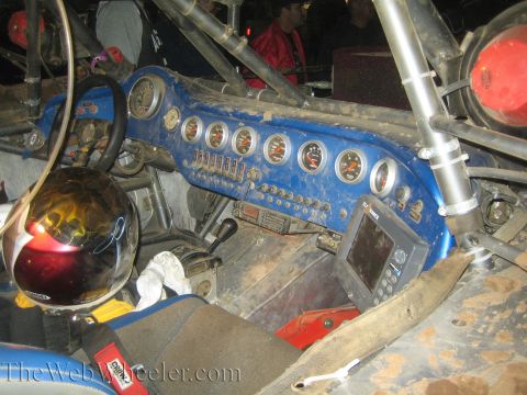 1990 Ford Bronco - cockpit of an off-road race truck shows a blue gauge panel with multiple dials, switches, a flame-graphic helmet and a GPS screen.