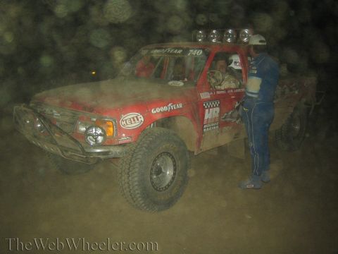 1990 Ford Bronco - driver in a blue suit stands beside a red number 740 off-road race truck with a roof light bar and Hella lights in a dusty night pit.