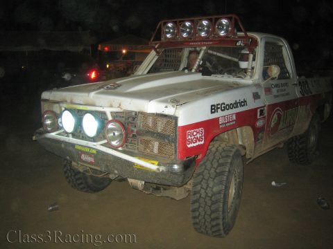 1990 Ford Bronco - white-and-red off-road race truck with a four-lamp roof bar and BFGoodrich livery is parked in a dusty night pit area.