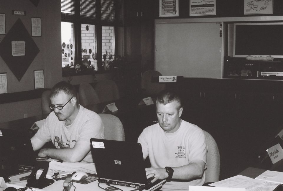 Two men sit at desks with laptops in a room with windows and framed items on the wall
