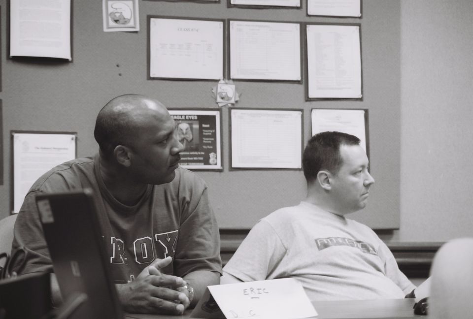 Two men sit at a table, one wearing a t-shirt with 'ROY' on it