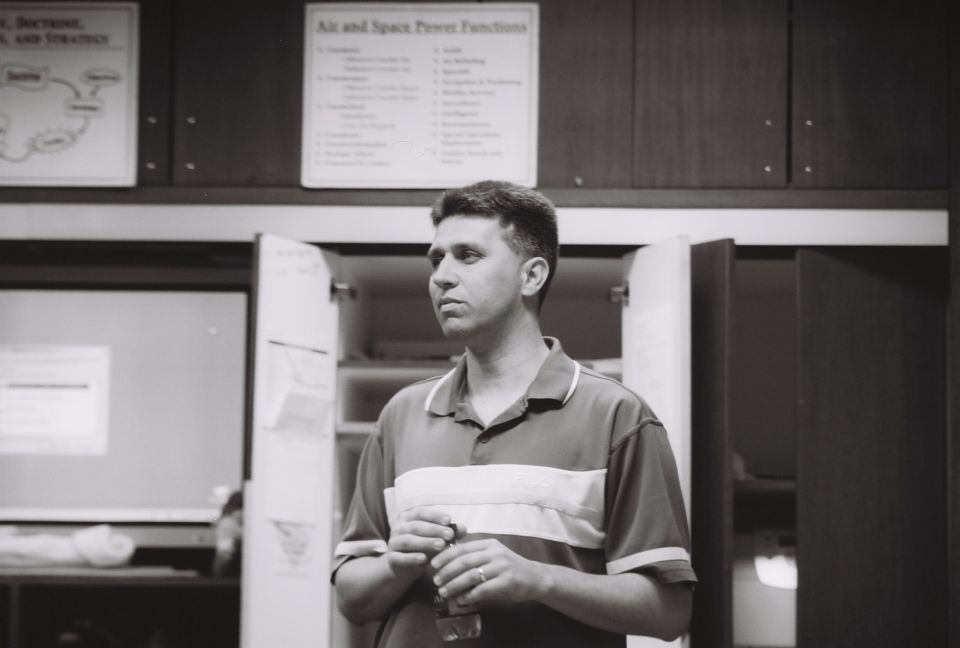Man in a collared shirt holding a bottle, standing in front of open cabinets with charts on the wall