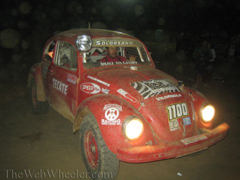 1990 Ford Bronco - mud-covered red VW Beetle Baja Bug race car, number 1100 and Tecate decals, runs with headlights on during a nighttime desert race.