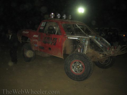 1990 Ford Bronco - red tube-frame off-road race truck, number 801 with twin upright exhaust stacks and a roof light bar, sits in the dark with a figure beside…