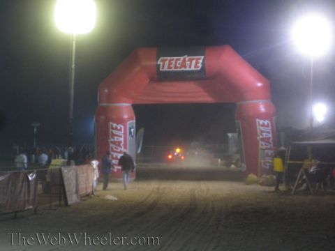 1990 Ford Bronco - red inflatable Tecate arch spans a dirt race course at night, lit by tall balloon lights with spectators and tents along the sides.