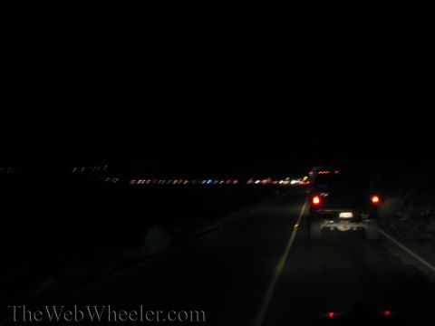 1990 Ford Bronco - blurry nighttime highway scene with a vehicle's red tail lights ahead and a band of distant city lights along the horizon.