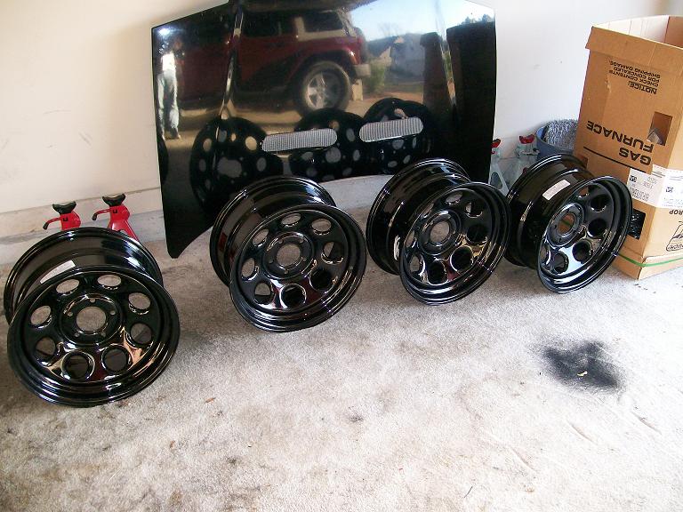 Four gloss black steel wheels on garage floor in front of a black hood reflecting a red Jeep