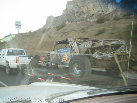 1990 Ford Bronco - Seen through a windshield, a blue tube-frame off-road buggy number 136 rides on a trailer towed by a white pickup near a rocky hillside.
