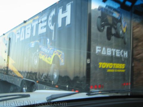 1990 Ford Bronco - side of a black Fabtech enclosed trailer showing a yellow race truck graphic and Toyo Tires logo, viewed through a windshield.