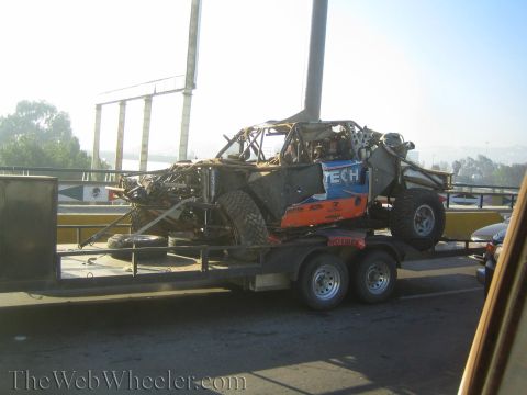 1990 Ford Bronco - wrecked tube-frame off-road race truck in blue and orange sits on a tandem-axle trailer on a highway in morning light.