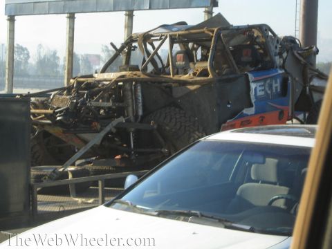 1990 Ford Bronco - stripped and damaged tube-frame off-road race truck on a trailer, photographed in traffic beside a white car under an overpass.