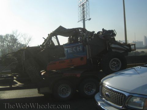 1990 Ford Bronco - wrecked blue and orange tube-frame off-road race truck rides on a tandem trailer towed by a silver Ford pickup on a highway.